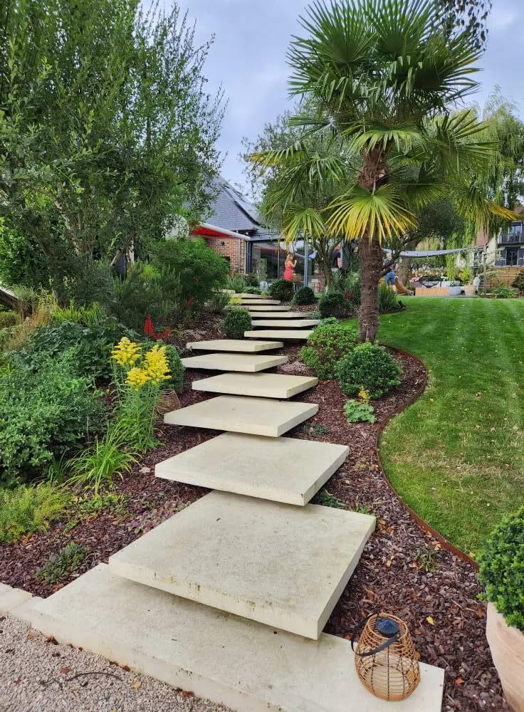 Allée en dalles béton flottantes traversant un jardin paysager avec palmiers et massifs fleuris, aménagement paysager résidentiel dans le Calvados