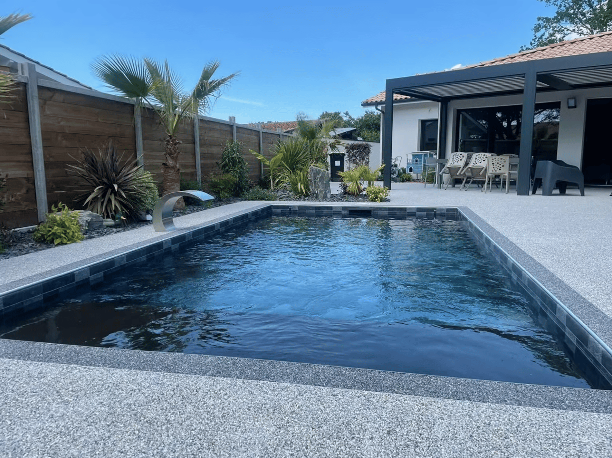 Piscine entourée d’une terrasse grise en moquette de pierre, palissade en bois et végétation