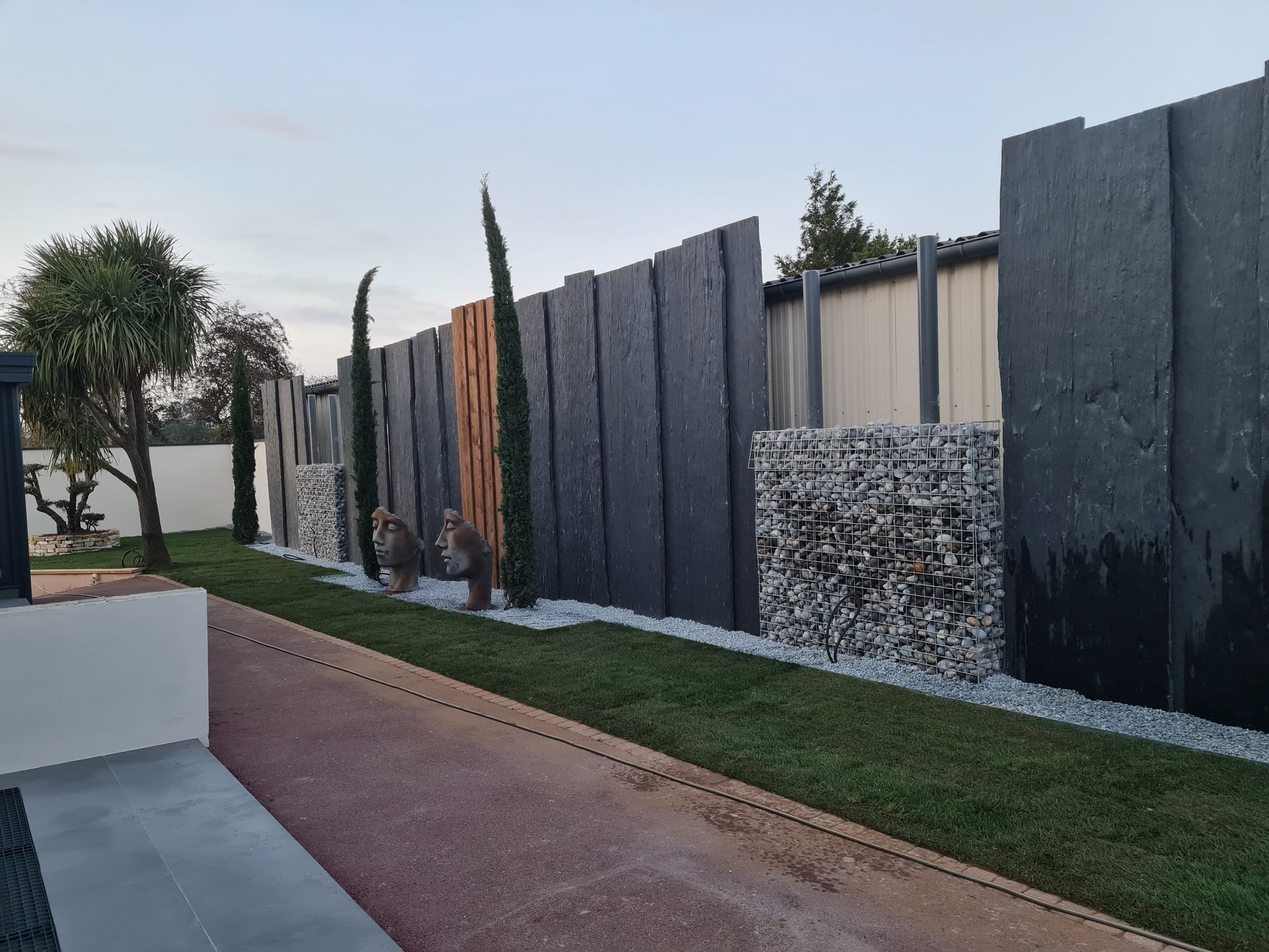 Clôture sombre avec bois, végétaux et un gabion dans un jardin