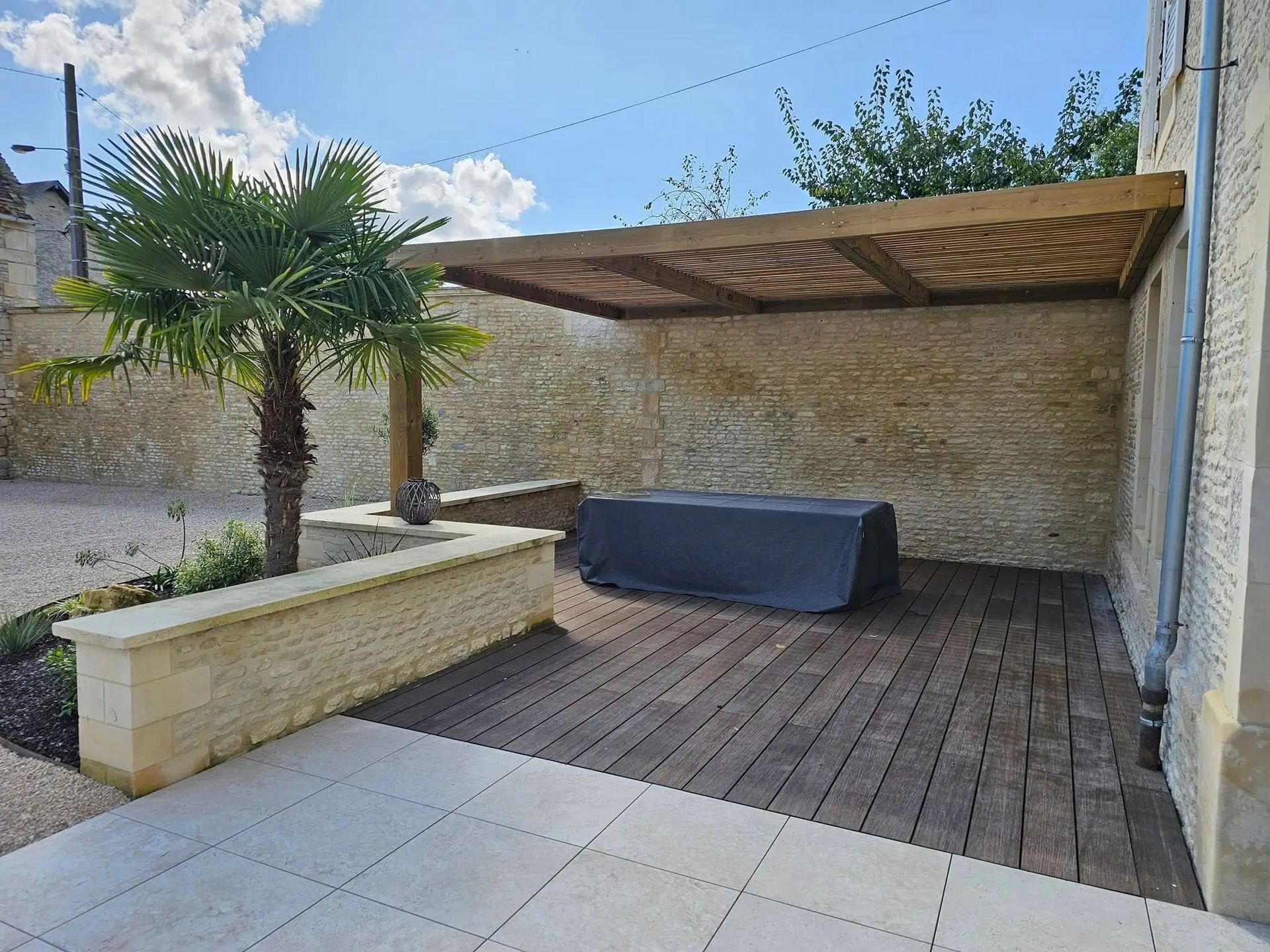Terrasse en bois avec pergola et palmier devant un mur en pierre